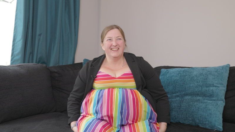 Steph, a local resident in Essex. Steph is sitting on a sofa and is wearing a rainbow striped dress.