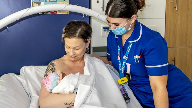 Women sitting in hospital bed breastfeeding her baby with Midwife looking on