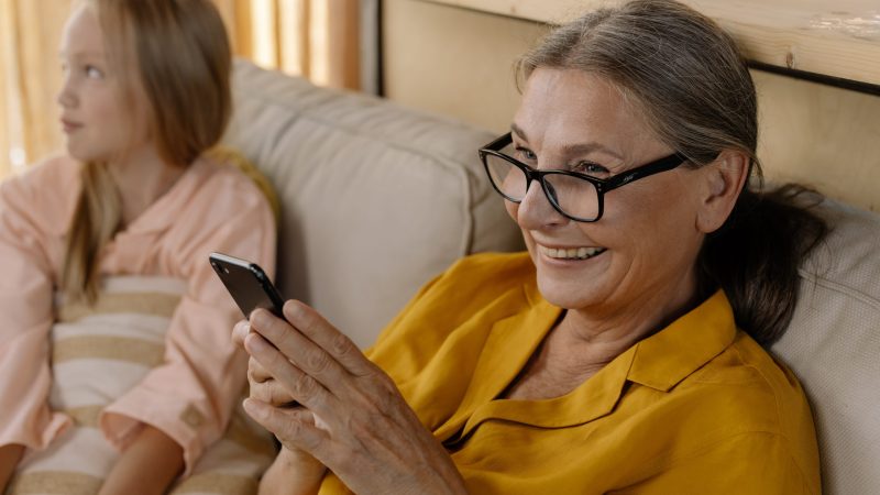 A girl sitting on a couch with an older lady who is looking at a mobile phone