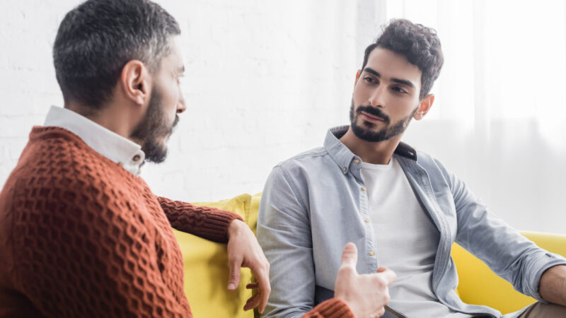 Hispanic son looking at father talking on blurred foreground on couch, two generations of men
