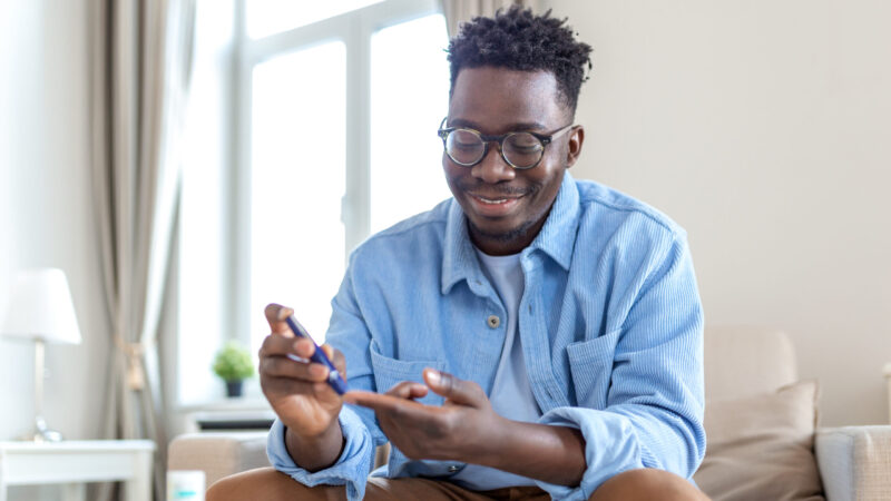 African man is sitting at the sofa at the home and taking blood from his finger due to diabetes. The daily life of a man of African-American ethnicity person with a chronic illness who is using glucose tester.