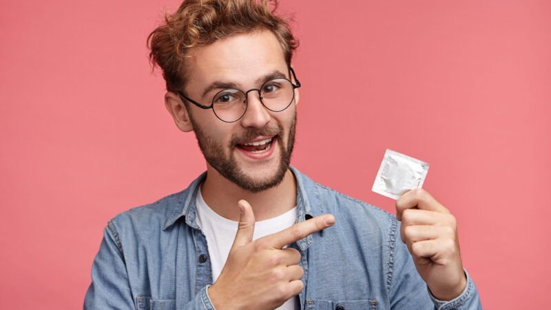 Handsome bearded man dressed casually, indicates at rubber condom, advertises new aromatic and qualified kind, recommends to buy it, guarantees good protection. Advertising concept. Contraception