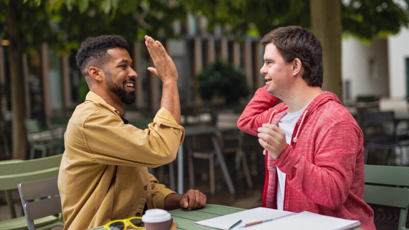 Young man with Down syndrome with mentoring friend sitting outdoors in cafe celebrating success.